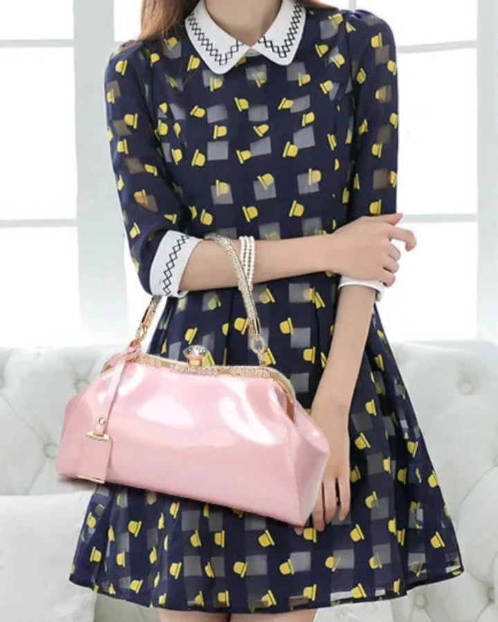 Woman wearing a navy dress with yellow patterns, holding a shiny pink patent handbag against a bright background.