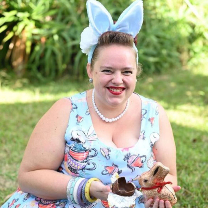 Woman wearing bunny ears and a whimsical dress, holding chocolate bunny in a garden setting.