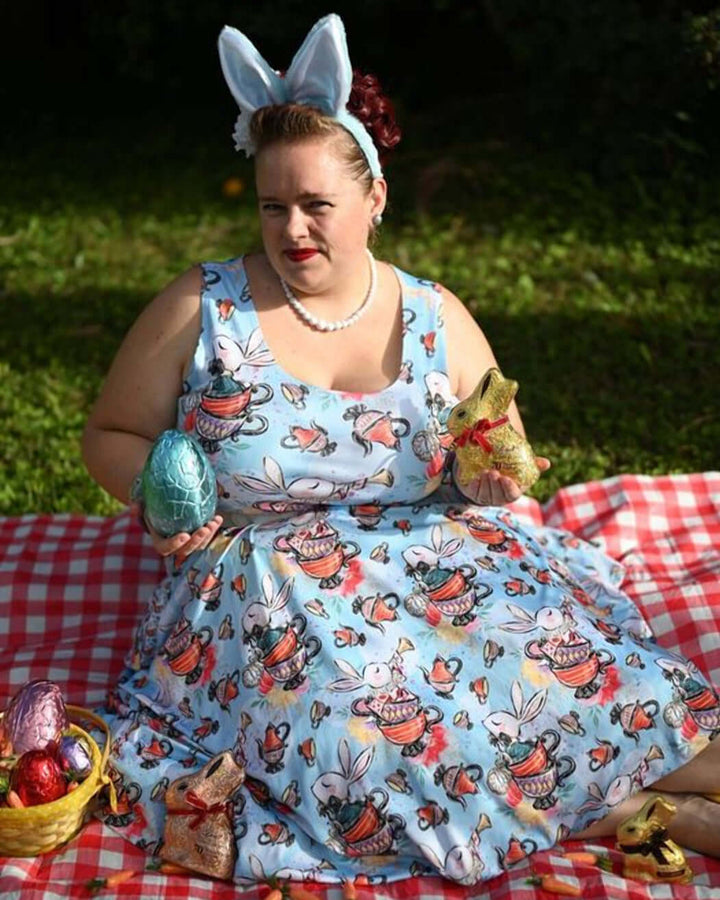 Woman in a blue floral skater dress with bunny ears, holding colorful Easter eggs on a picnic blanket.